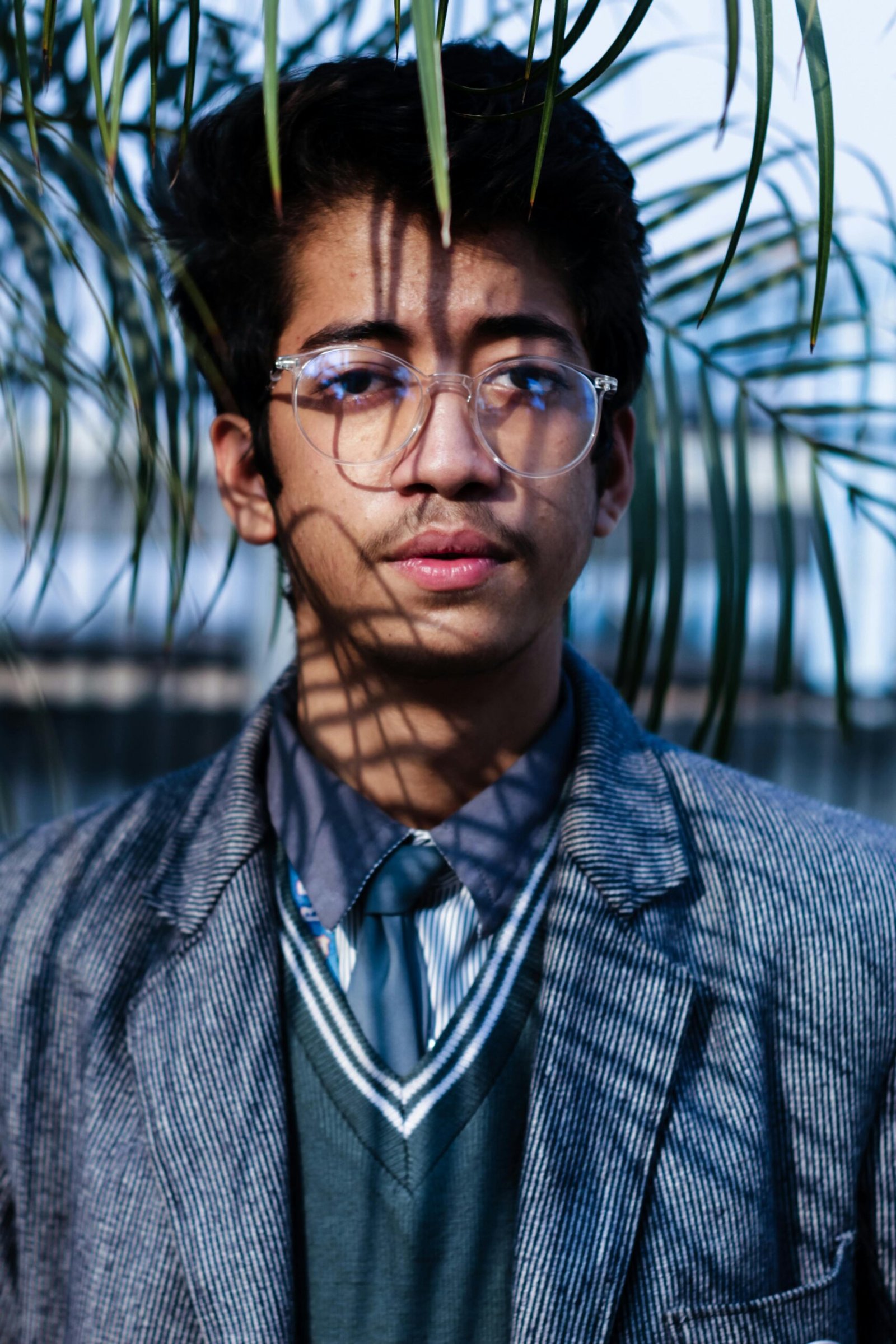 Portrait of a young man wearing eyeglasses and a suit in a natural setting with palm leaves.